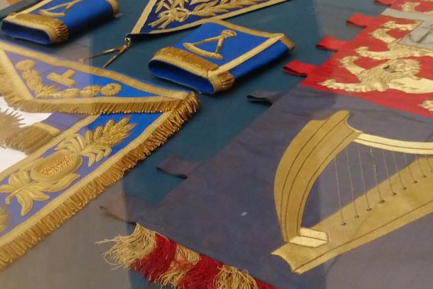 Gold, red and blue embroidered banner and masonc apron and gloves, laying flat in a glass display case